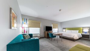 A modern hotel room with a teal sofa, a green ottoman, a desk with a computer, a bed with white bedding, and a television.