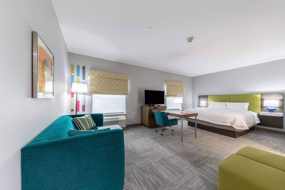 A modern hotel room with a teal sofa, a green ottoman, a desk with a computer, a bed with white bedding, and a television.