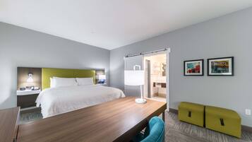 A modern hotel room with a bed, bedside table, lamp, and a desk with a chair. There are two framed pictures on the wall.
