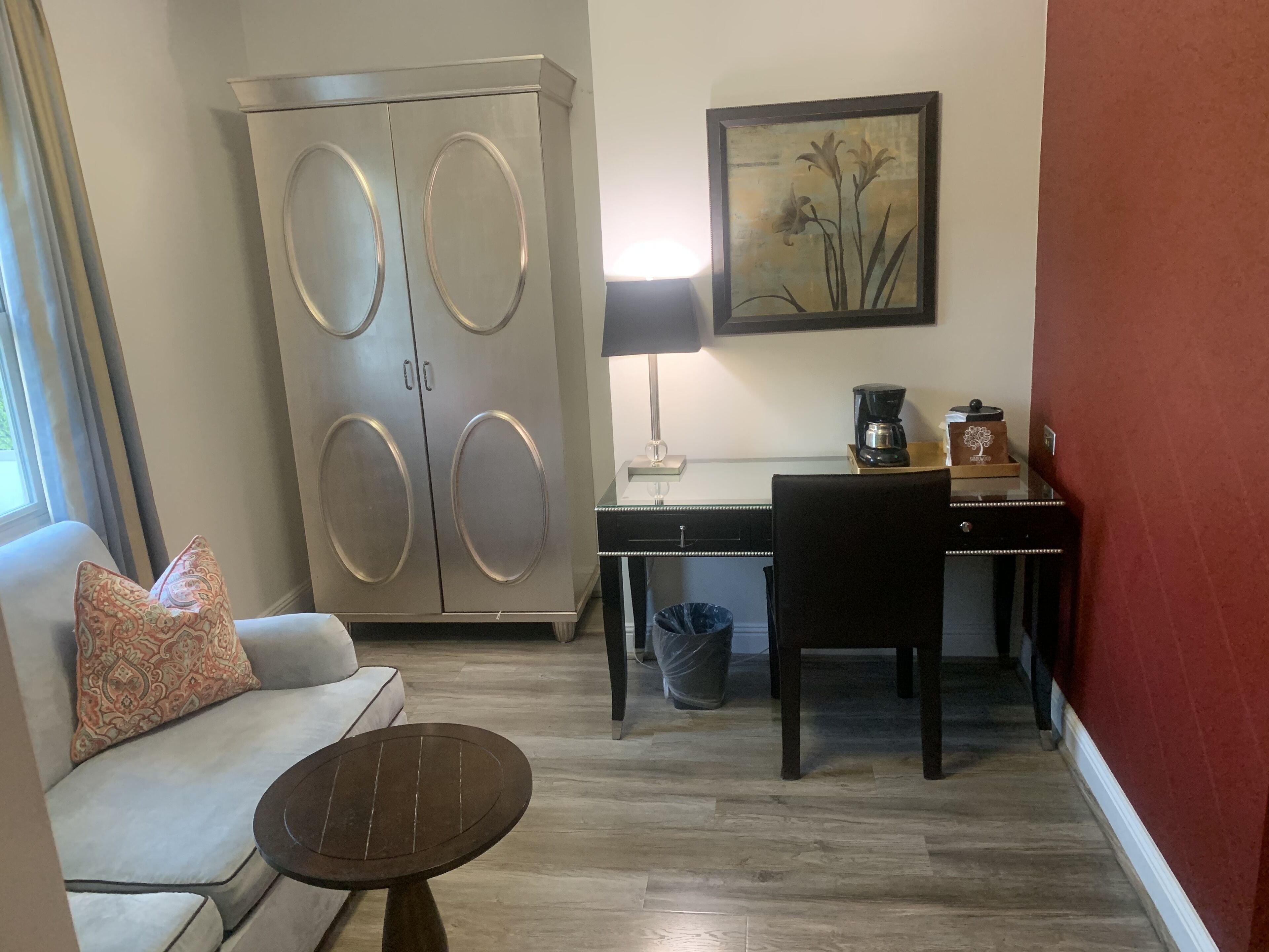 A compact hotel room with a sofa, a small round table, a desk with a lamp, a coffee maker, and a trash bin.