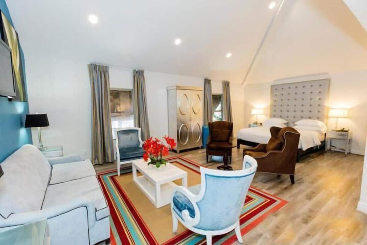A modern hotel room with a sofa, armchair, coffee table, and a bed with a headboard.