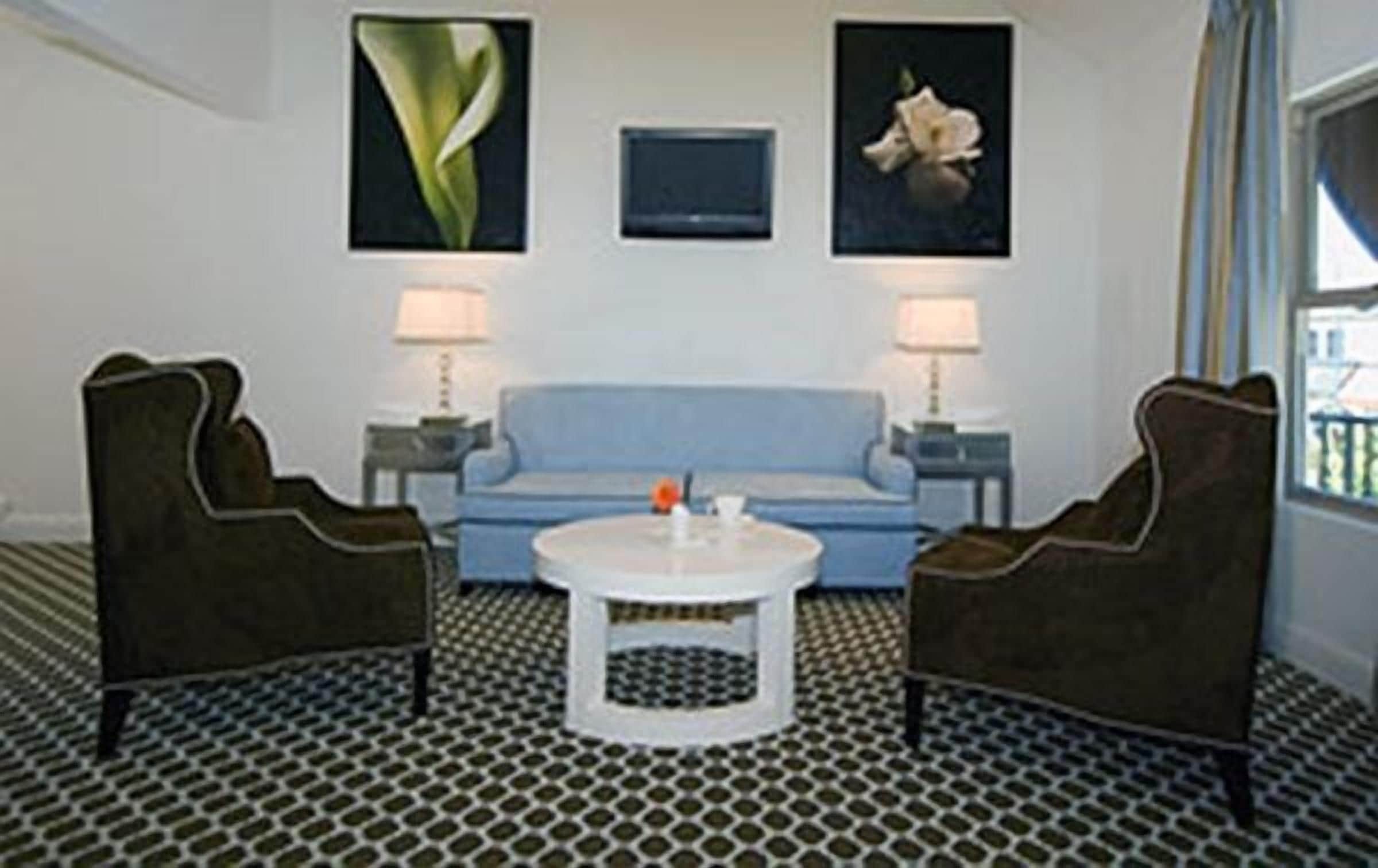 Une pièce avec un canapé bleu, deux fauteuils marron, une table basse blanche et un sol à motifs. Deux lampes murales et deux tableaux encadrés sont accrochés au mur au-dessus du canapé.