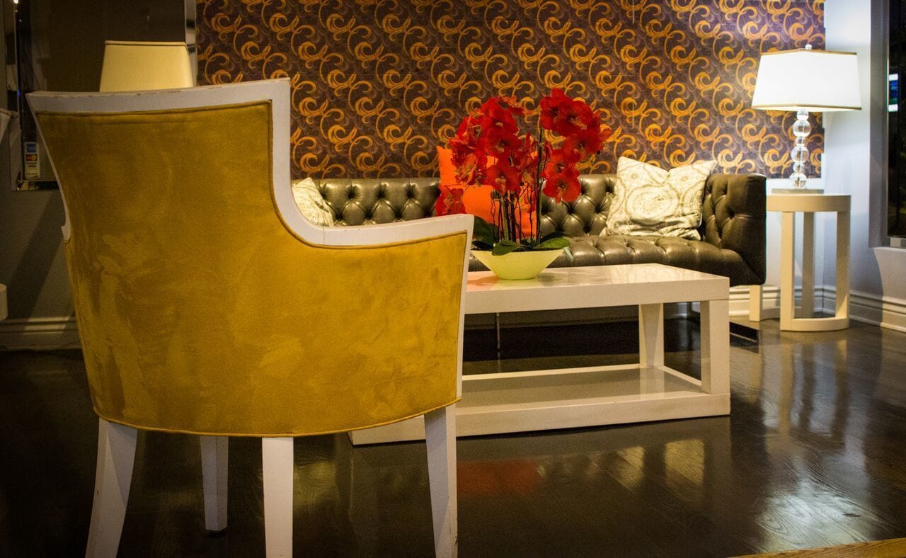 A room with a yellow armchair, a tufted leather sofa, a white coffee table, and a patterned wallpaper background.