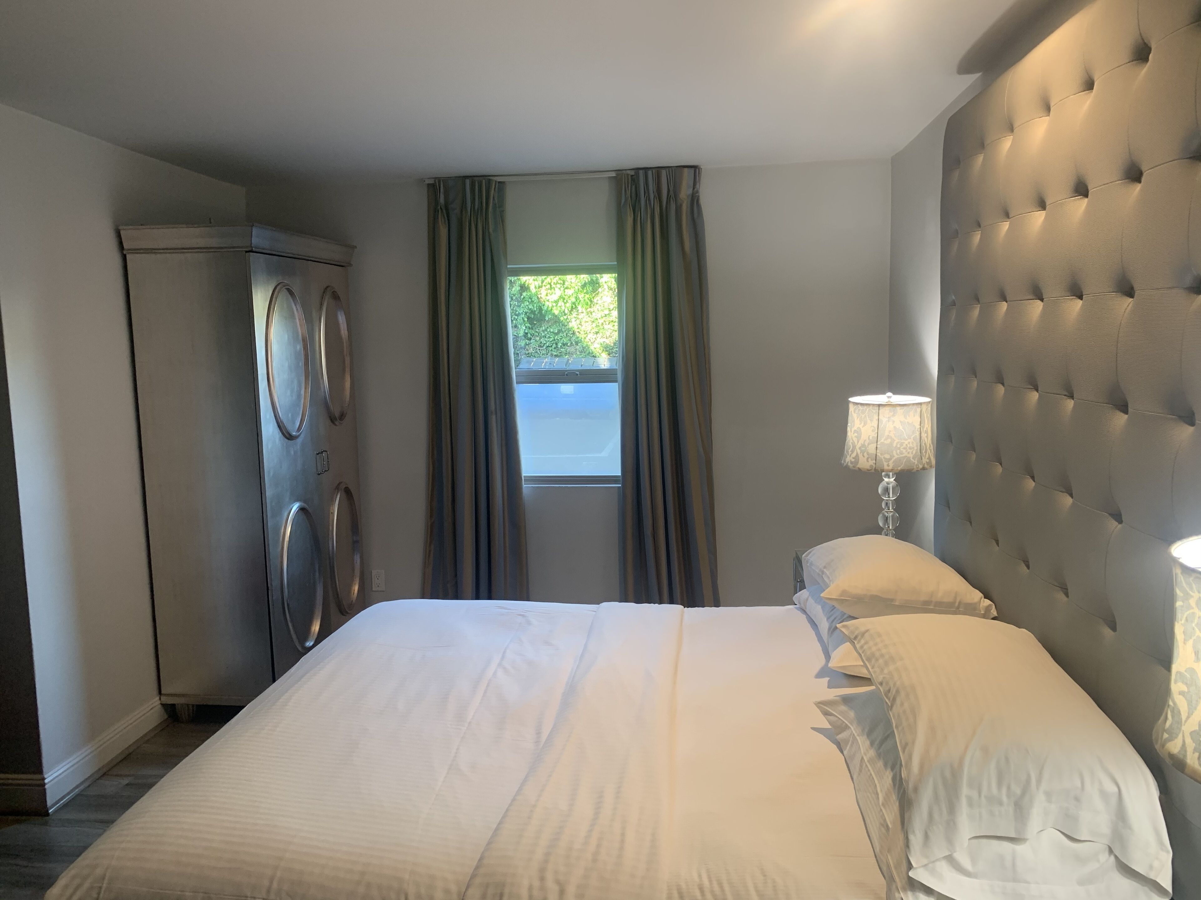 A hotel room with a large bed, a headboard, a window with curtains, a lamp, and a washing machine.