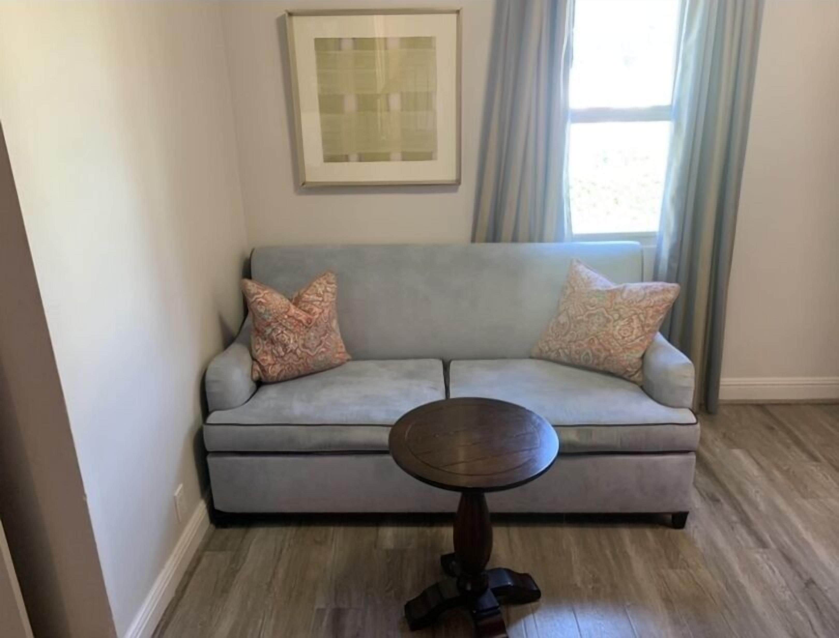 A living room with a grey sofa, two patterned cushions, a round wooden coffee table, and a framed picture on the wall.
