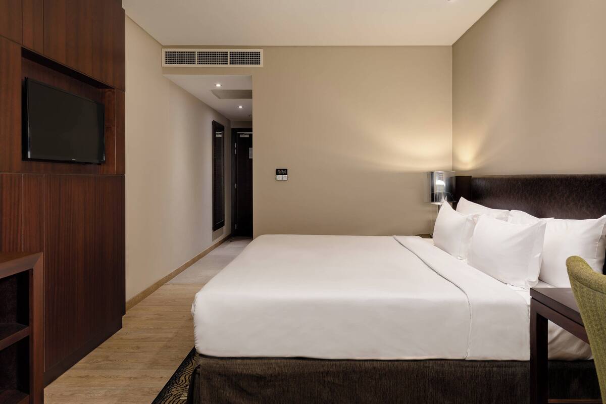 A hotel room with a large bed, a television mounted on a wooden panel, a bedside table with a lamp, and a door leading to another room.
