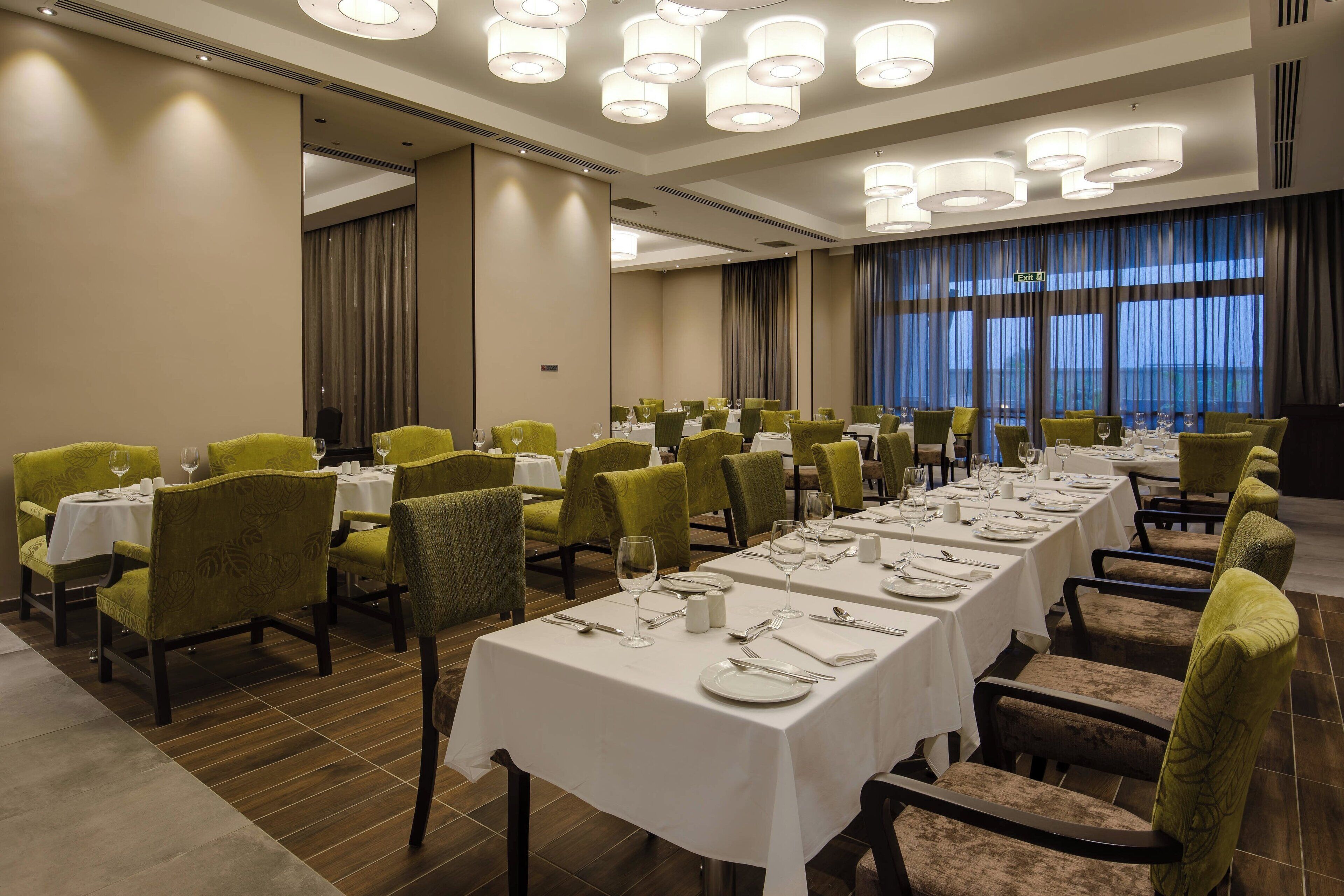 A conference room with multiple tables set for dining, green upholstered chairs, and a large window with curtains.