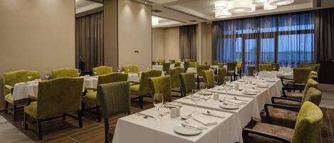 Un salón de conferencias con varias mesas dispuestas para comer, sillas tapizadas de verde y un amplio ventanal con cortinas.