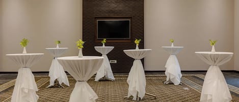 A conference room with six round tables, each with a white tablecloth and a small plant. The room has a carpeted floor and a large screen on the back wall.