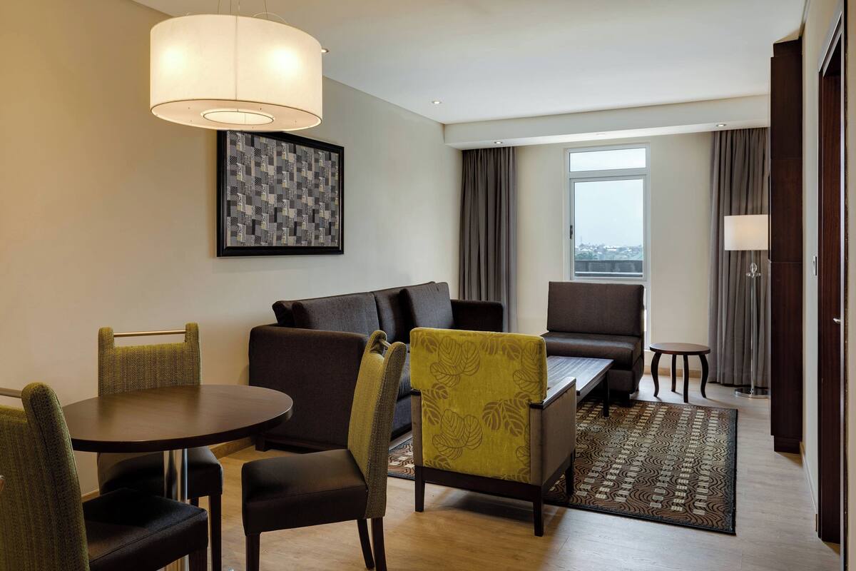 A modern hotel room with a dining area, a sofa, a patterned rug, and a large window.