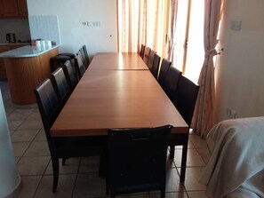 Una taula de dinar llarga amb caires negres, una zona de cuina amb armaris de fusta i una finestra amb cortines.