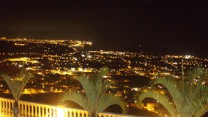 Un paisaje urbano nocturno con vistas a edificios iluminados y un balcón con palmeras.