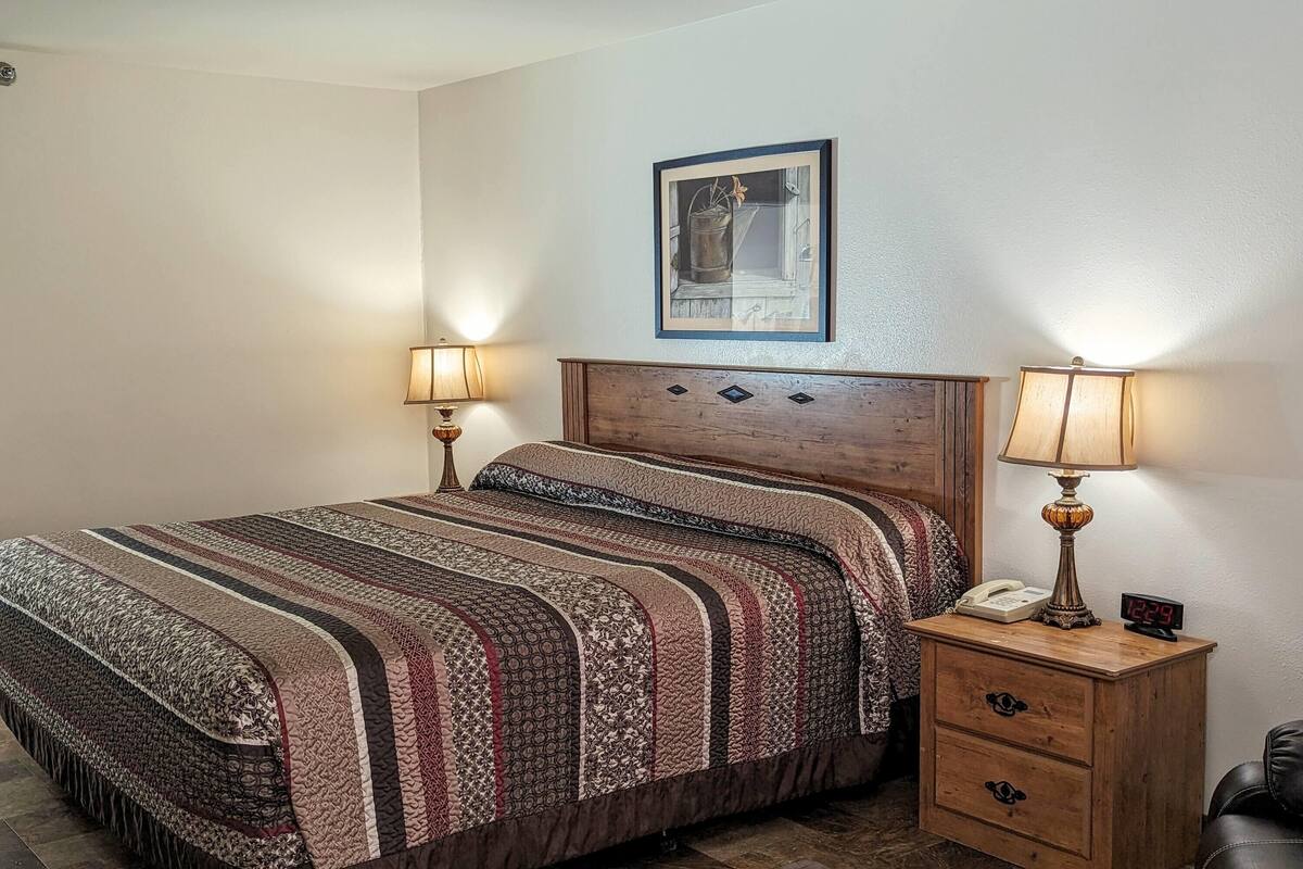 A bedroom with a bed, two bedside lamps, a nightstand with a phone and clock, and a framed picture on the wall.