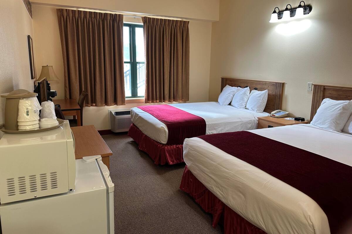 A hotel room with two beds, a desk with a lamp, a small refrigerator, and a window with curtains.
