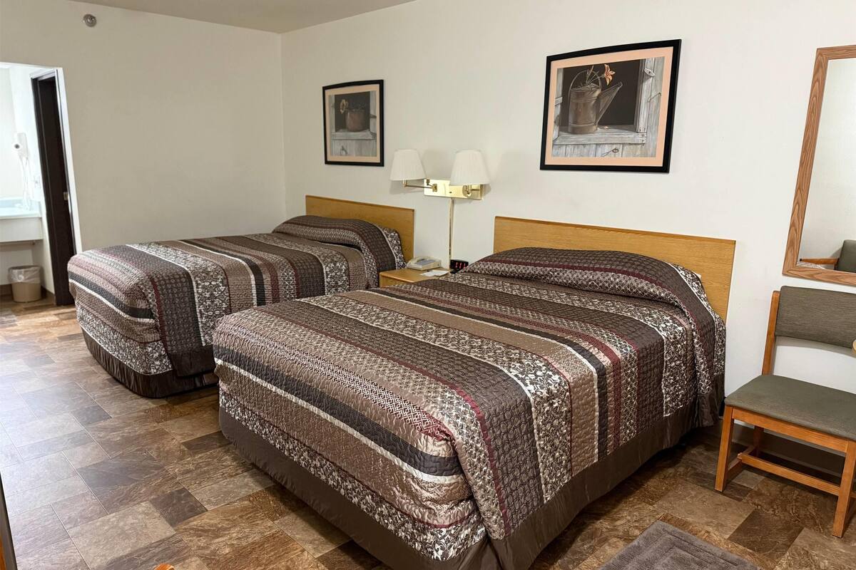 A hotel room with two beds, a nightstand, a lamp, and a chair.