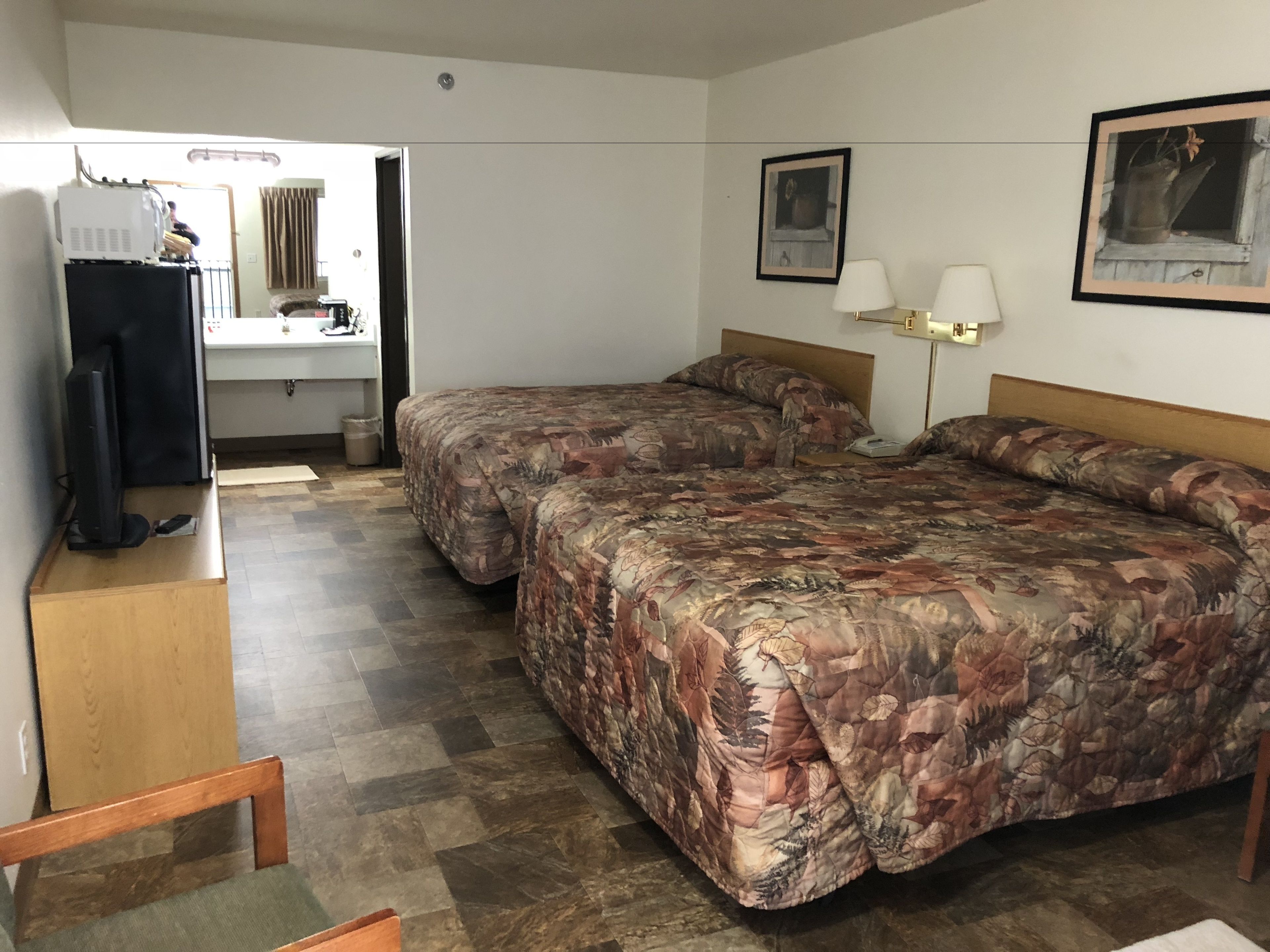 Habitación de hotel con dos camas, televisión, escritorio y baño visible a través de una puerta abierta.