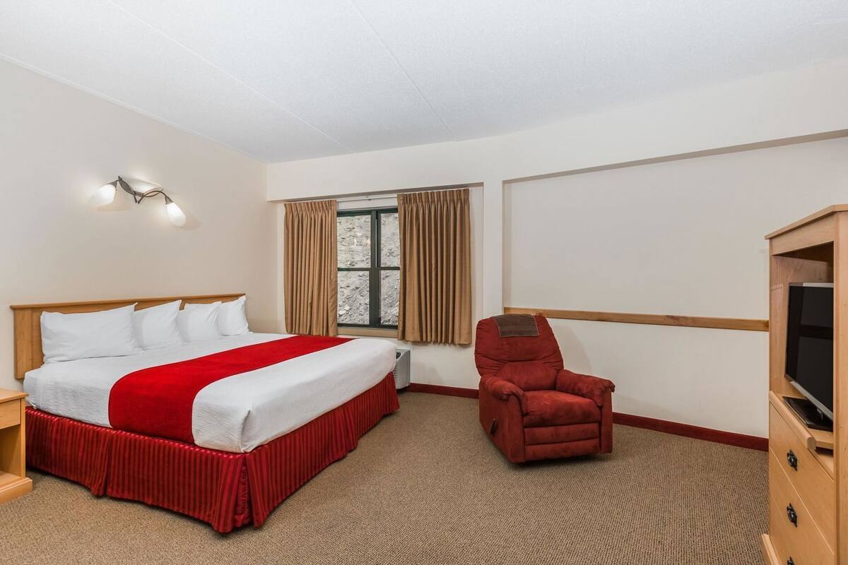 A hotel room with a bed, a red armchair, a television, and a window with curtains.
