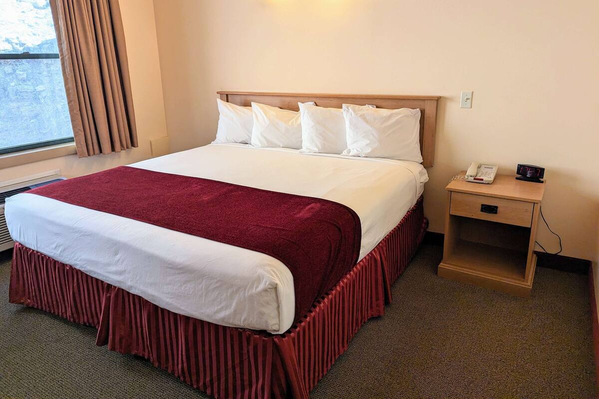 A hotel room with a large bed, a wooden headboard, a bedside table with a phone and alarm clock, and a window with curtains.