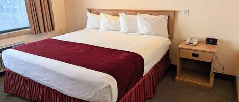 A hotel room with a large bed, a wooden headboard, a bedside table with a phone and alarm clock, and a window with curtains.