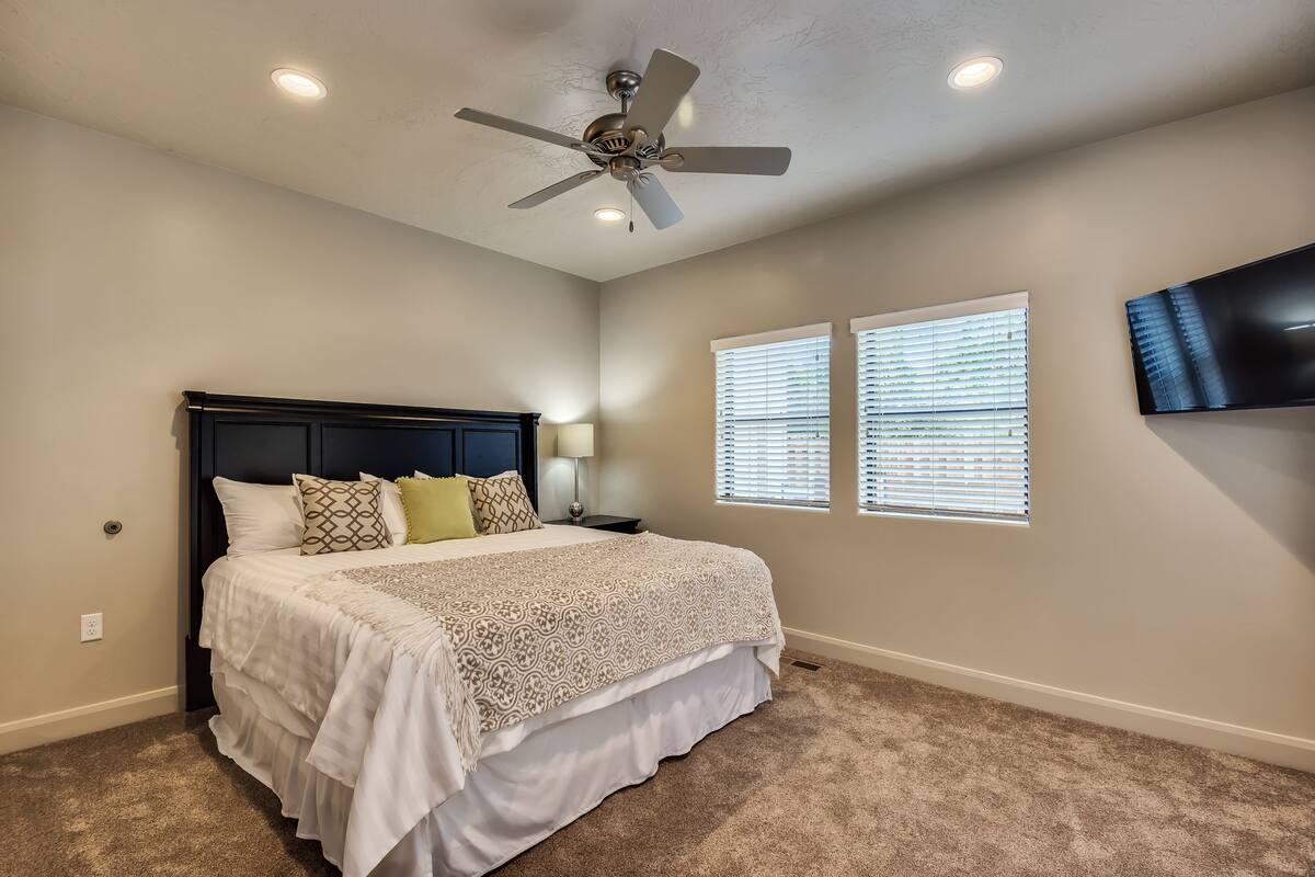 A bedroom with a bed, ceiling fan, two windows with blinds, and a TV mounted on the wall.