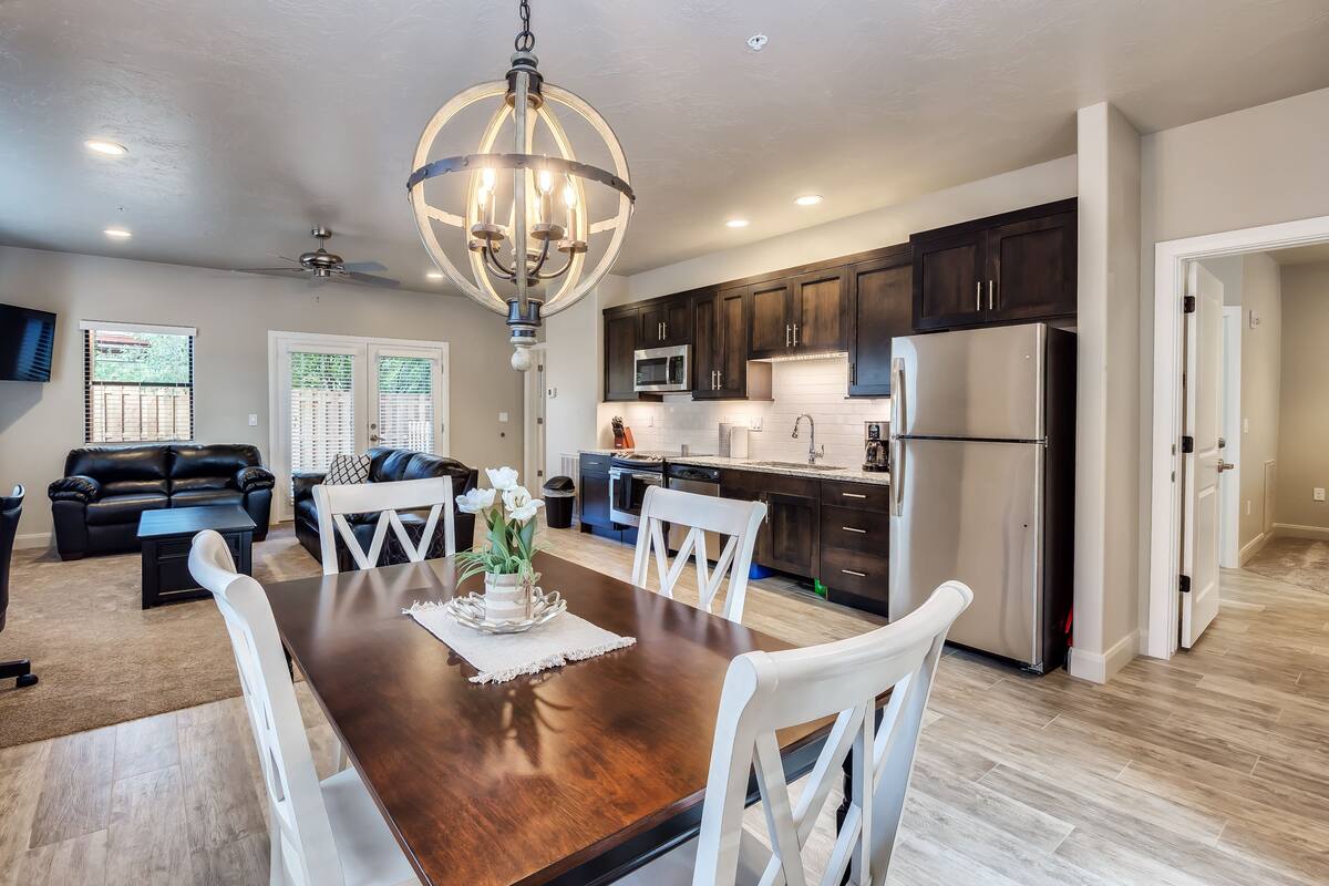 A spacious living room with a dark wood dining table, white chairs, and a chandelier. There is a kitchen area with dark cabinets and stainless steel appliances.