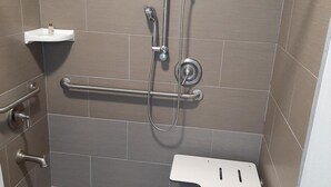 Un área de ducha pequeña y accesible con barra de apoyo, cabezal de ducha de mano y estante blanco.