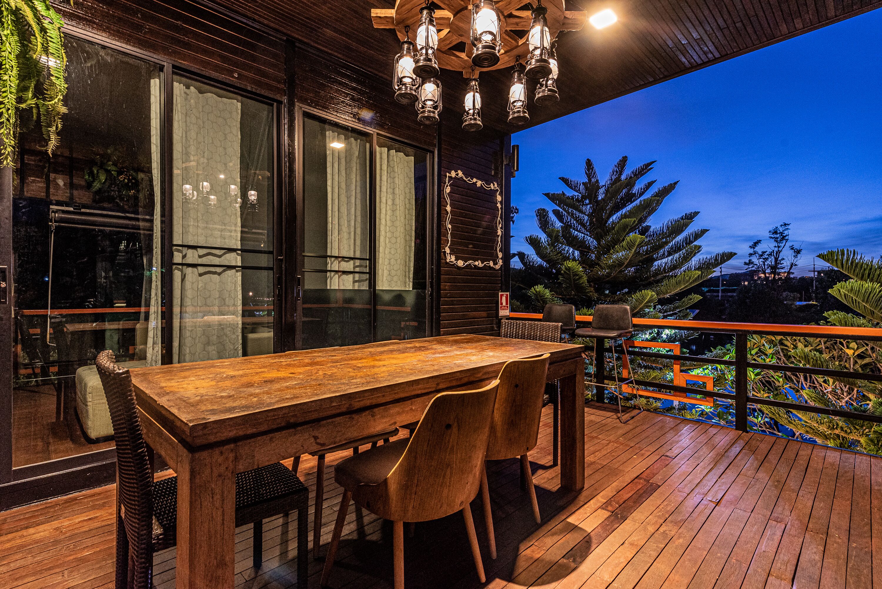 A wooden deck with a dining table and chairs, large windows, and a view of the outdoors.