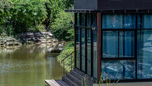 Um deck de madeira com escadas que levam a uma estrutura envidraçada à beira de um lago tranquilo, cercada por vegetação exuberante.