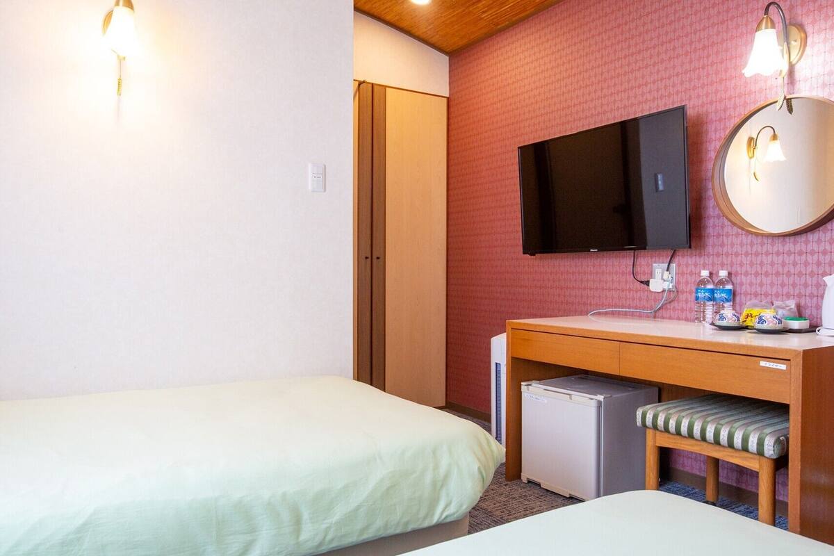 A hotel room with two single beds, a small desk with a mirror, a television, and a refrigerator.
