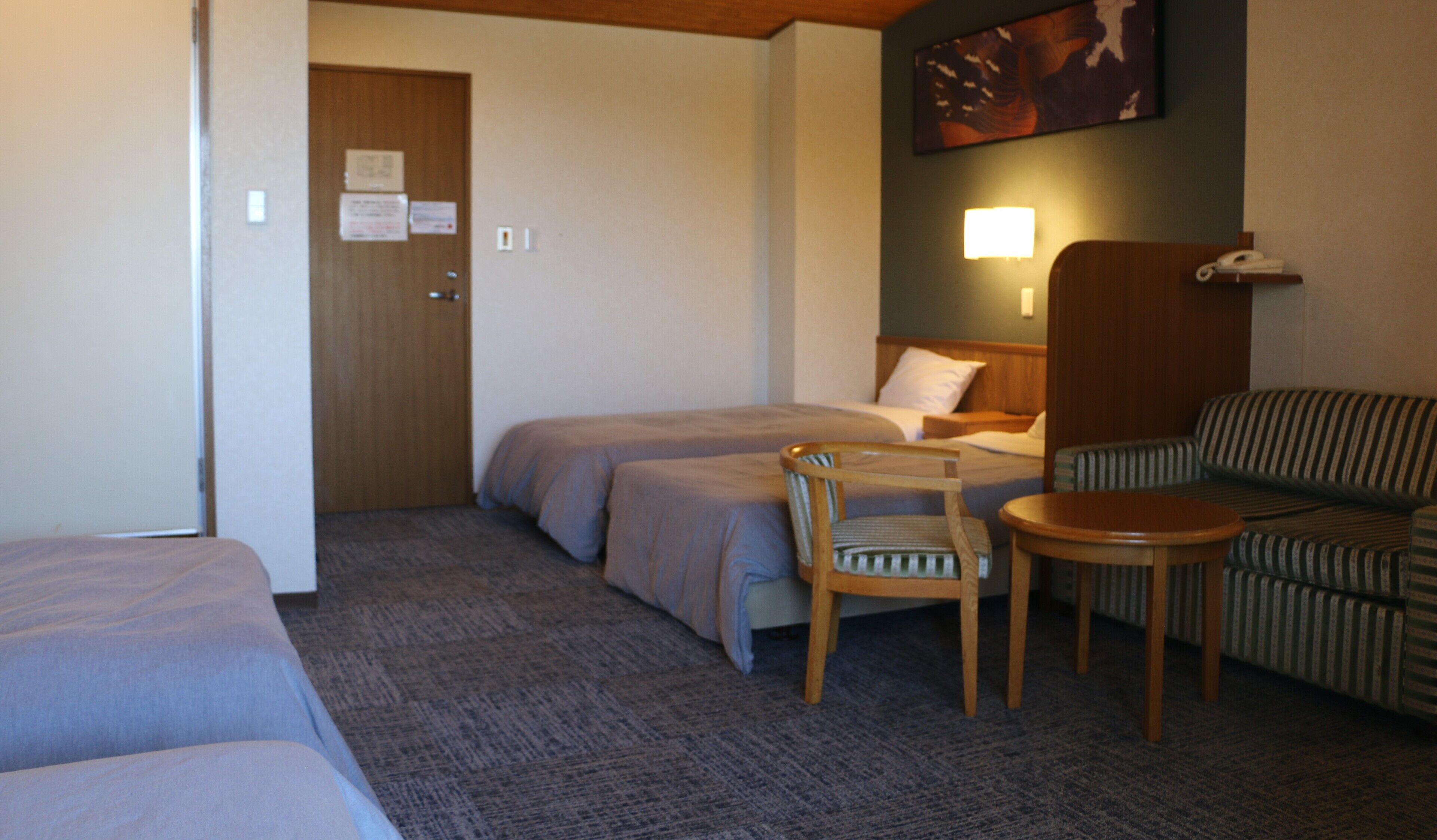 A hotel room with two single beds, a wooden chair, a striped sofa, a small table, and a painting on the wall.
