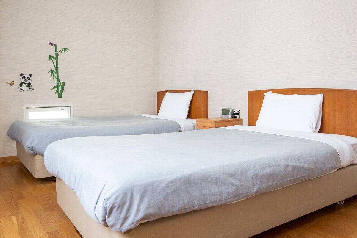 A hotel room with two single beds, wooden headboards, a bedside table with a clock, and a wall decoration of bamboo and a panda.