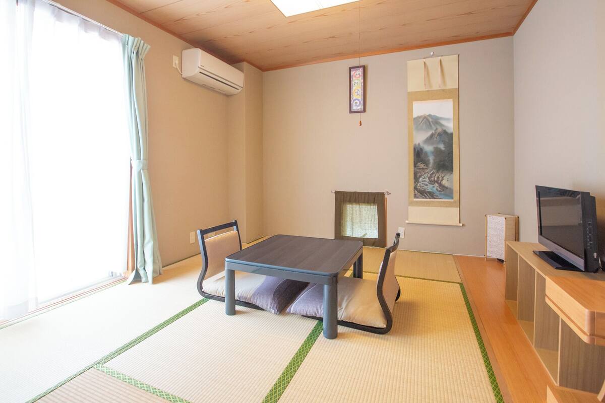 A room with tatami flooring, a low table with cushions, a television, and artwork on the walls.