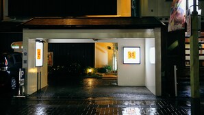 Entrada de hotel à noite, com calçada molhada, letreiros iluminados e um carro estacionado.