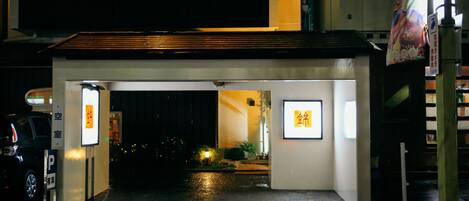 Ingresso di un hotel di notte, con marciapiede bagnato, insegne illuminate e un'auto parcheggiata.