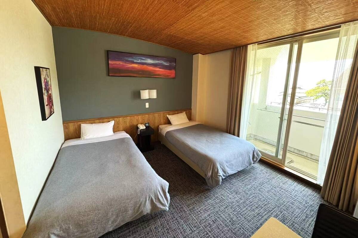 A hotel room with two single beds, a wooden ceiling, a painting on the wall, a small table with a lamp, and a large window with curtains.