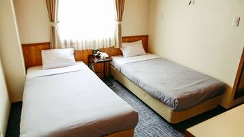 A hotel room with two single beds, a small table with a lamp, and a window with curtains.