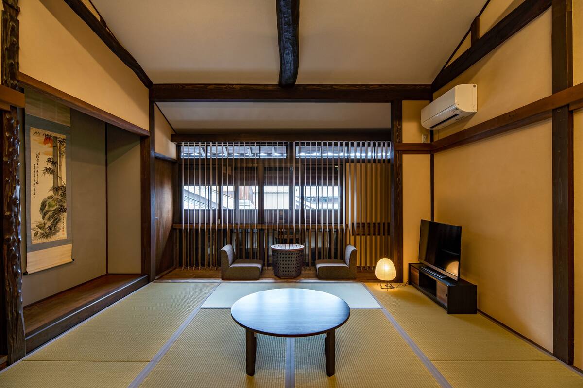 Una habitación tradicional japonesa con suelo de tatami, una mesa baja, dos sillas acolchadas, un televisor y una ventana con persianas verticales.