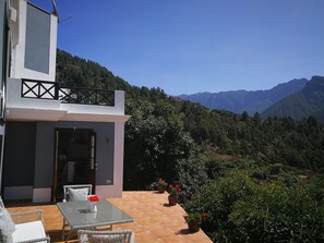 Edificio moderno con un patio que tiene una mesa y sillas, rodeado de macetas con plantas y vistas a un paisaje montañoso.