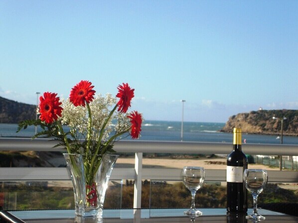 Una mesa con un jarrón de flores rojas y una botella de vino, con vistas a la costa.