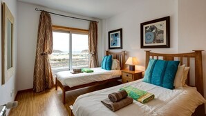 Een hotelkamer met twee bedden, een raam met gordijnen, een nachtkastje met een lamp en kunstwerken in een lijst aan de muur.