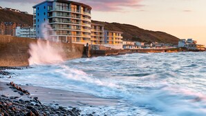 Un hotel frente al mar con varios balcones que dan al océano, un muro de piedra frente a la costa y una playa rocosa.