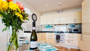 Una cocina moderna con armarios blancos, un microondas y una tetera. Hay una mesa de comedor con una botella de vino y un florero.