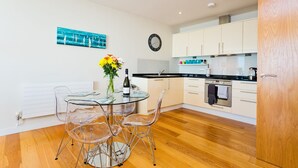 Una cocina moderna con armarios blancos, encimera negra y suelo de madera. Hay una mesa de comedor con sillas transparentes y un jarrón con flores.