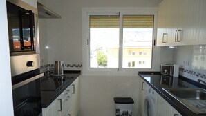 A compact kitchen with white cabinets, a black countertop, a window with blinds, a washing machine, and a trash bin. - Apartment With Stunning Sea Views And Vision Of The Rock Of 🇬🇮Gibraltar (Estepona)