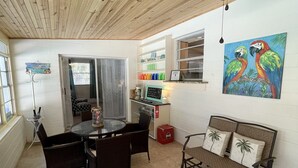A cozy patio area with a round table and chairs, a cushioned bench with palm tree pillows, a painting of colorful parrots, and a ceiling fan. - Walk to Beach! Family Beach House, Game room!  Sunset View! (Daytona Beach)