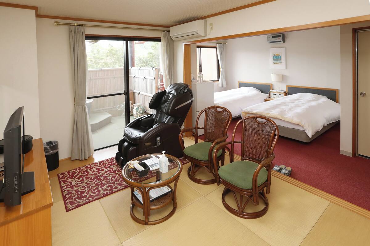 A hotel room with a massage chair, two green cushioned chairs, a bed with white bedding, a TV, and a small table with a remote control and a bottle.