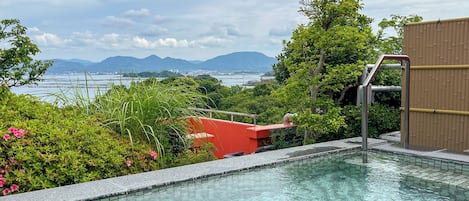 A swimming pool with a view of the sea and mountains, surrounded by greenery and a red building.