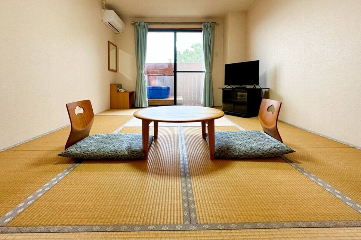A room with tatami flooring, a round table, two chairs, a television, and a sliding door leading to an outdoor area.