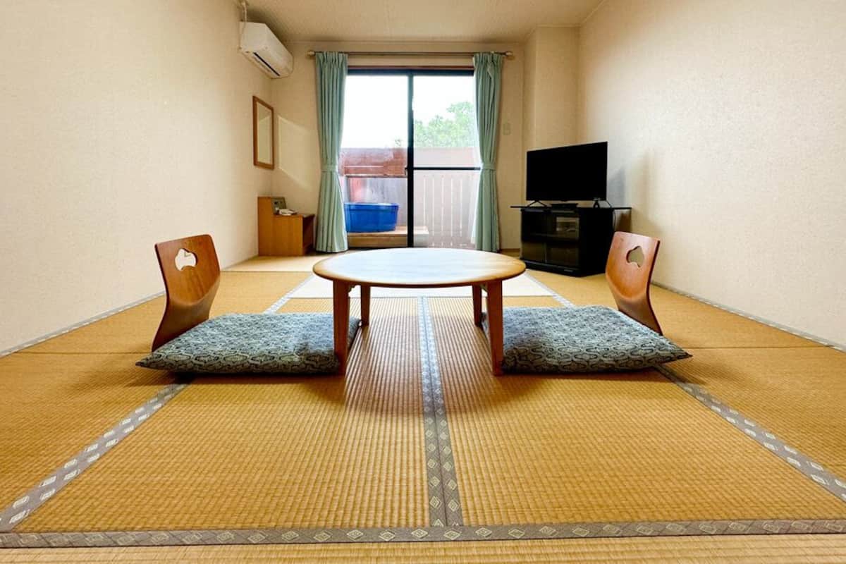 Una habitación con suelo de tatami, una mesa redonda, dos sillas, un televisor y una puerta corredera que da a un espacio al aire libre.