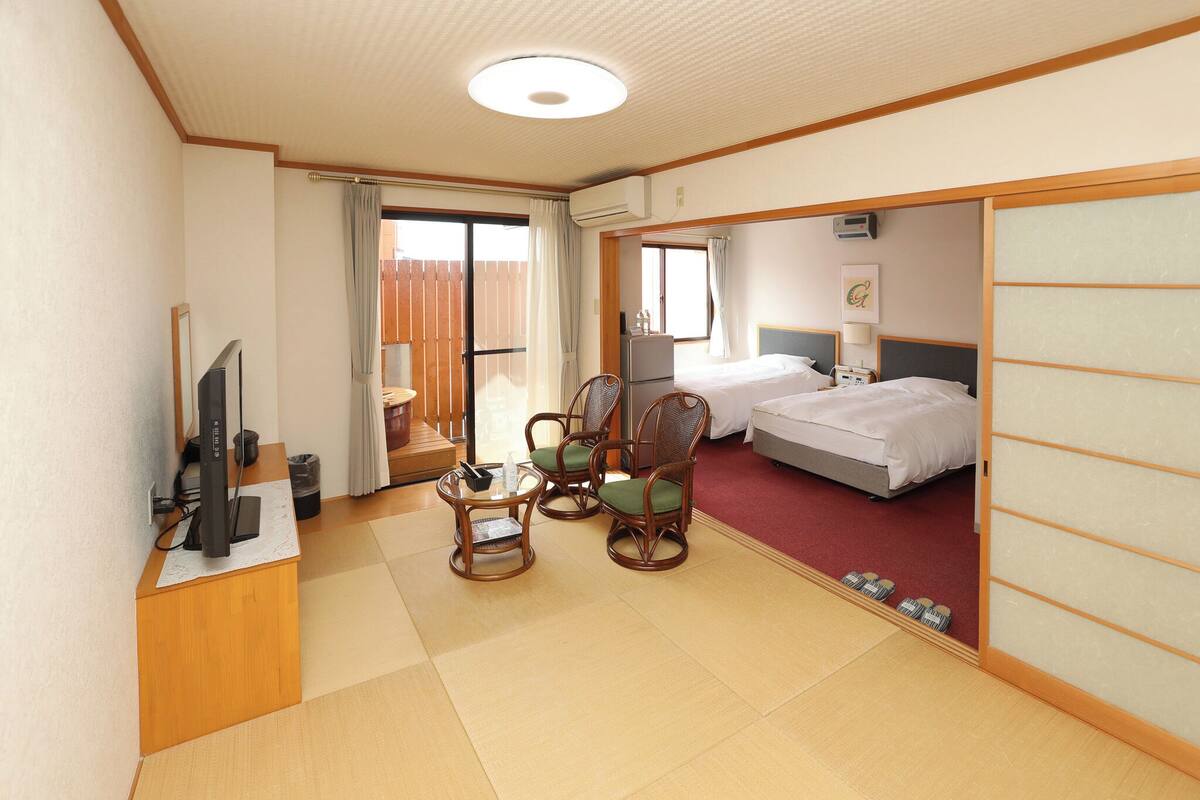 A hotel room with a bed, a desk with a computer, two chairs, and a sliding door leading to a balcony.